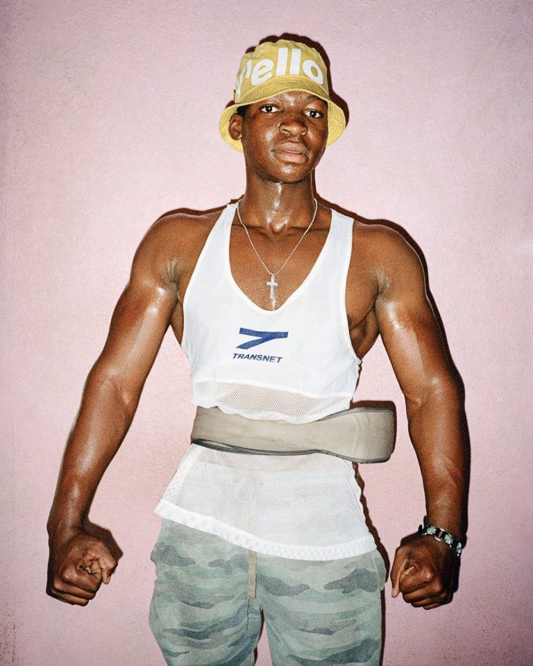 Dainius Ščiuka fotografija vaizduojanti sportišką jauną vaikiną dėvintį sportinį diržą, baltus sportinius marškinėlius ir geltoną kepurę rožiniame fone